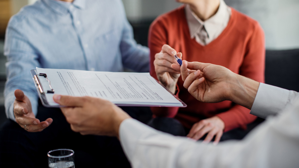 Close-up of woman signing an agreement with insurance agent while being on a meeting with her husband.